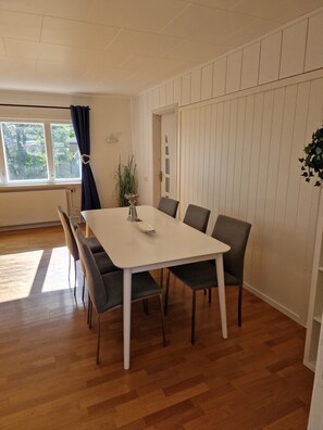 Een eethoek met een witte tafel en grijze stoelen, een plant in pot en een raam met gordijnen.
