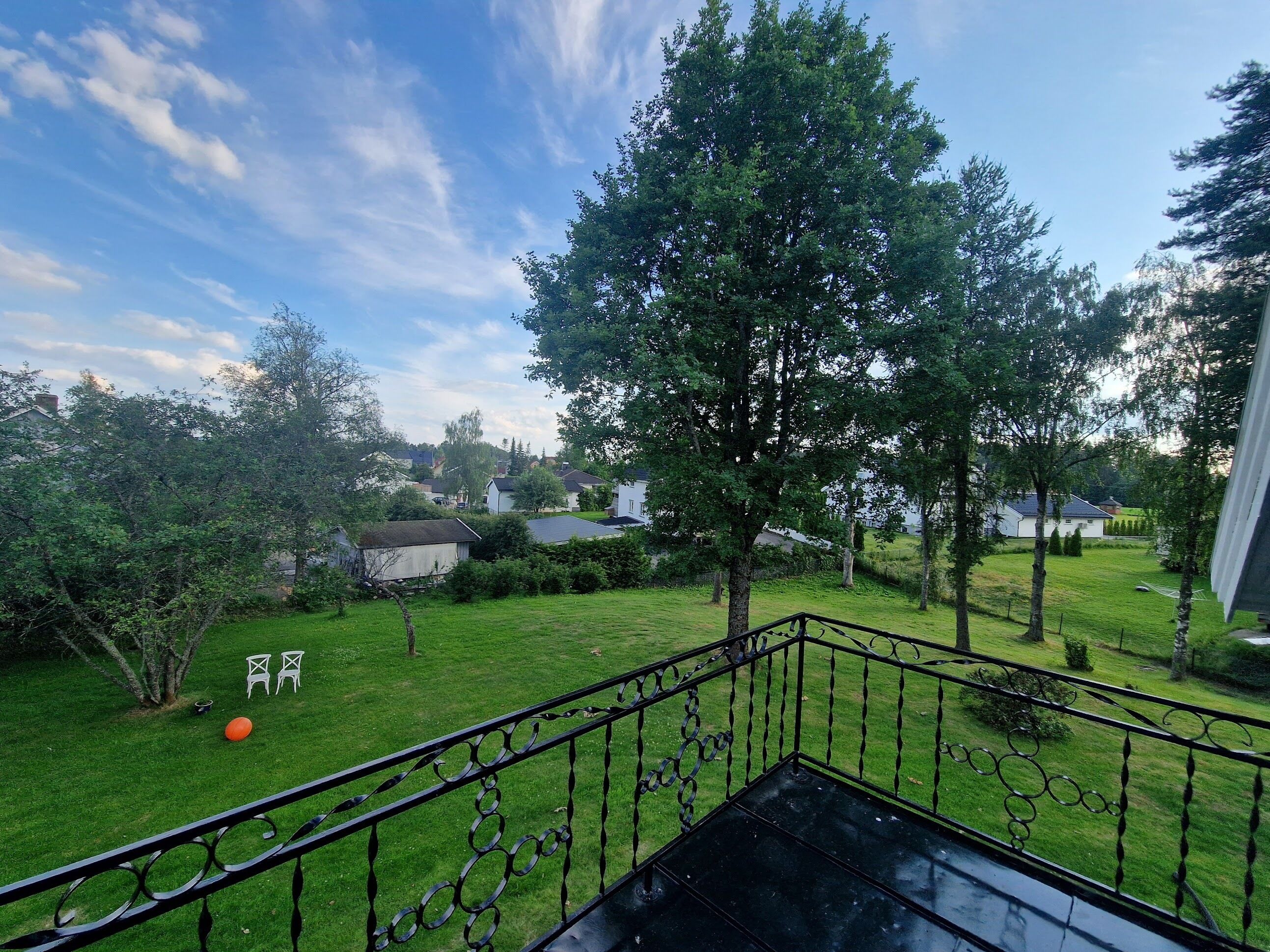 A balcony with a metal railing overlooking a grassy yard with trees and houses in the background.