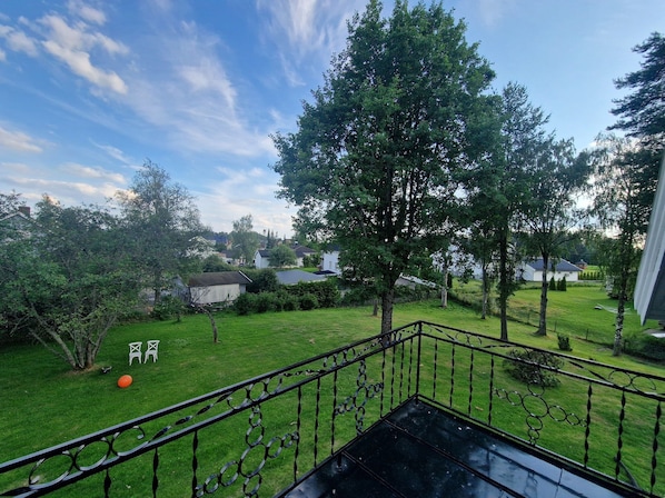 A balcony with a metal railing overlooking a grassy yard with trees and houses in the background.