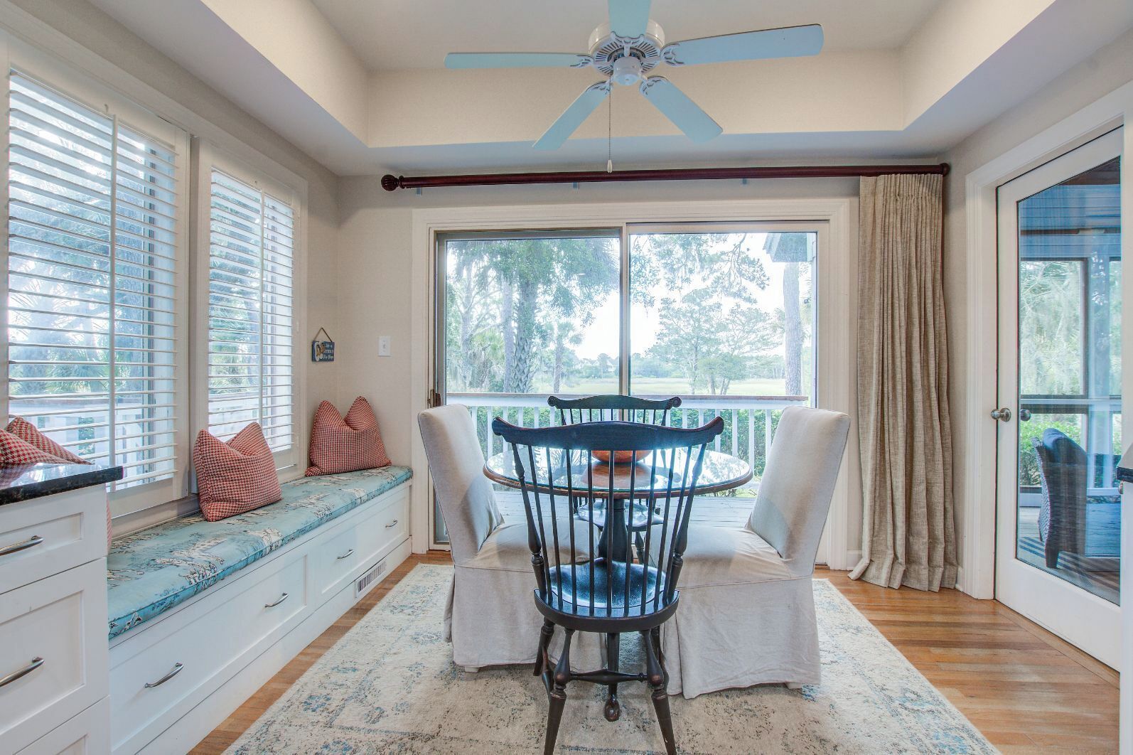 Une pièce comprenant une table à manger, des chaises, un banc avec des coussins, un ventilateur de plafond, de grandes fenêtres et une vue sur l’extérieur.