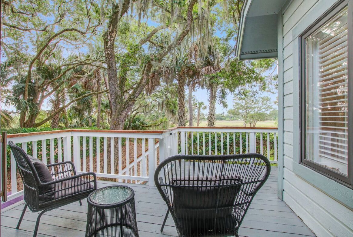 A covered outdoor area with two wicker chairs and a small table, surrounded by white railings and large trees with hanging moss.