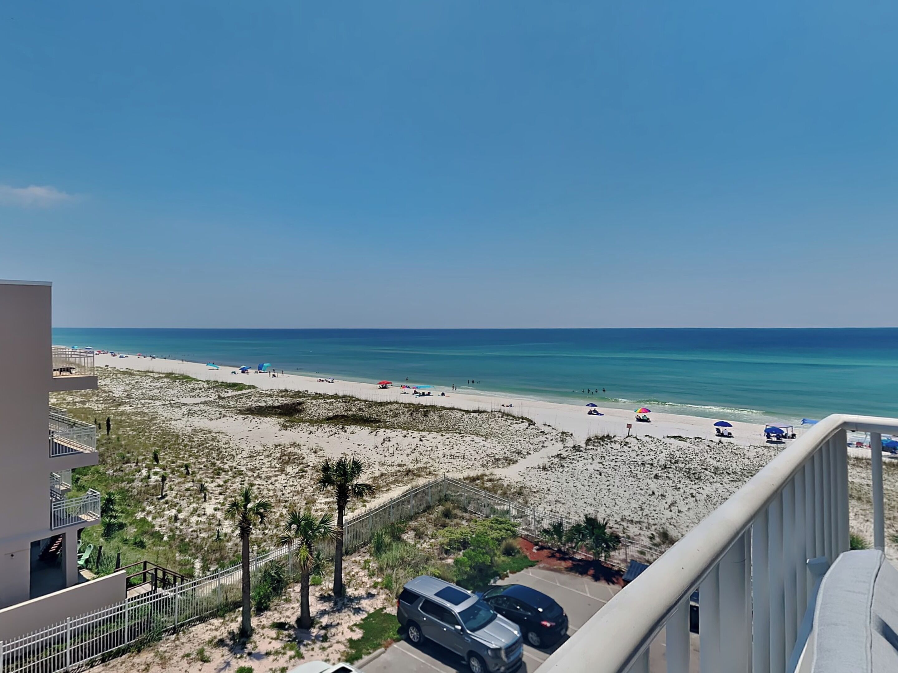 Blick auf einen Strand vom Balkon aus, mit blauem Himmel, Sandstrand und einigen parkenden Autos.