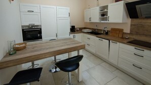 Cocina compacta con armarios blancos, encimera de madera y horno empotrado. Hay dos taburetes negros y una mesa pequeña.