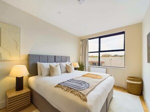 A hotel room with a bed, bedside table, lamp, window, and artwork on the wall.