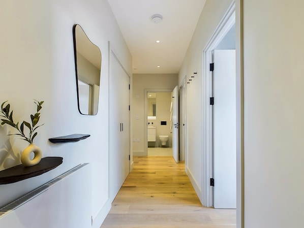 A hallway with a wooden floor, white walls, a mirror, and a plant on a shelf.