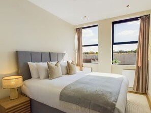 Habitación de hotel con una cama grande, una mesita de noche con lámpara y vistas a la ciudad a través de amplios ventanales.