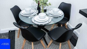 A round glass table with white plates, black chairs, and a vase with green leaves.