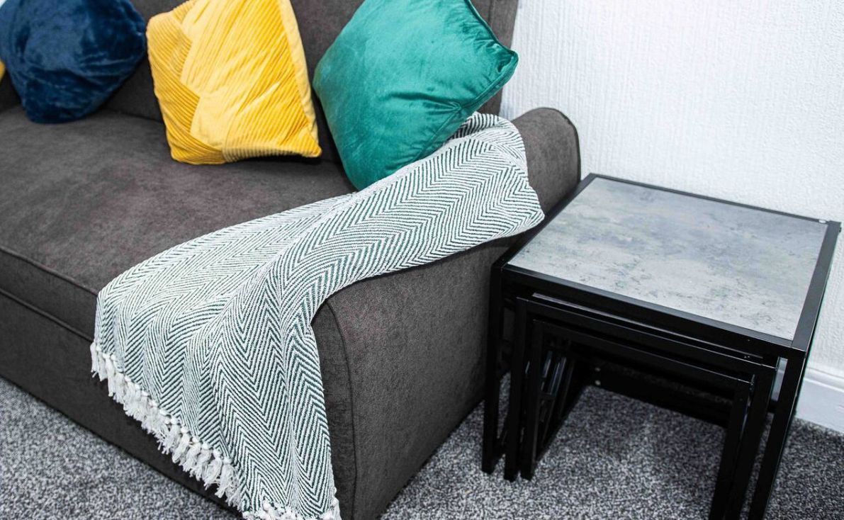 Un sofá gris con una manta de diseño, una mesita auxiliar negra y almohadas coloridas.