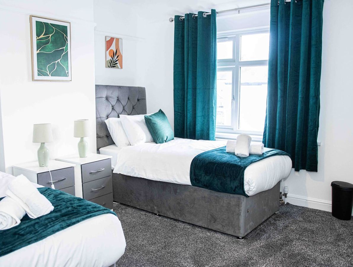 Habitación de hotel con dos camas, cabecero gris, cortinas color esmeralda y obras de arte en las paredes.
