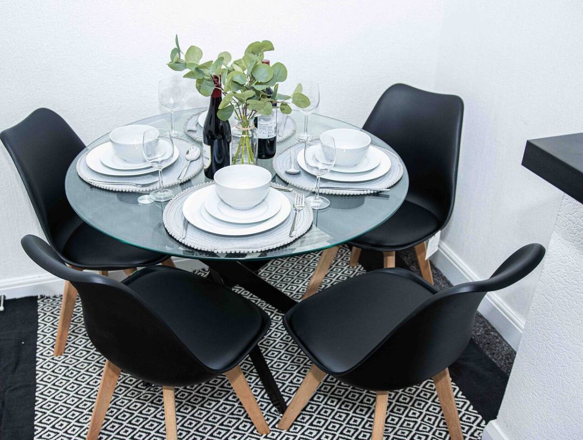 A round glass table with white plates, black chairs, and a vase with green leaves.