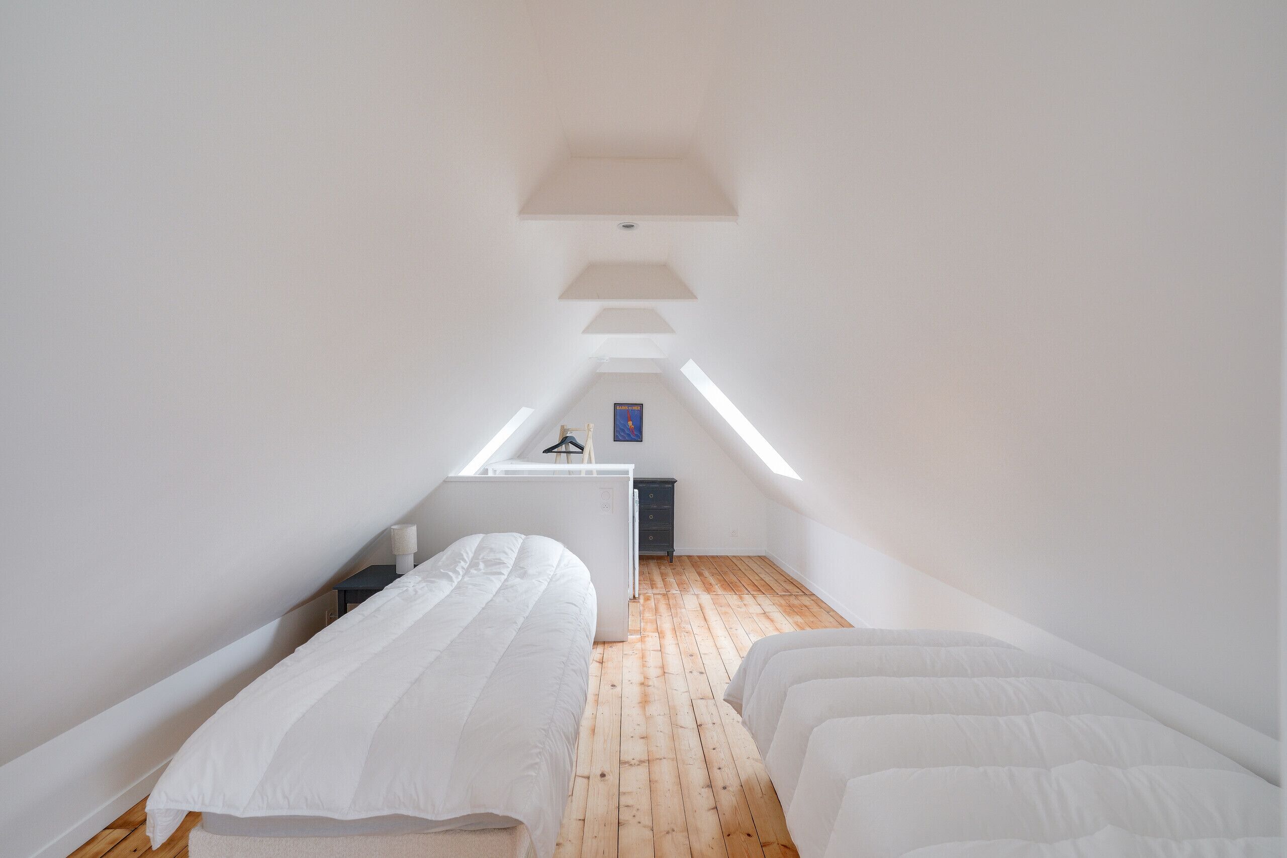 Une petite chambre mansardée aux murs blancs, avec deux lits, un bureau et une chaise.