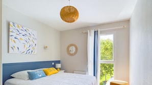 Een slaapkamer met een groot bed, een hanglamp, een raam met gordijnen, een klein tafeltje en een spiegel aan de muur.