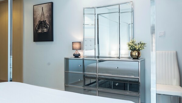 A modern hotel room with a large bed, a glass-fronted dresser, a bedside lamp, a framed picture, and a white chair.