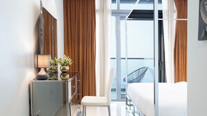 A modern hotel room with a bed, a desk with a lamp, a chair, a small table with a vase, and a large window with curtains. - Rooftop pool-2BR Apartment Near Downtown (Dubai)
