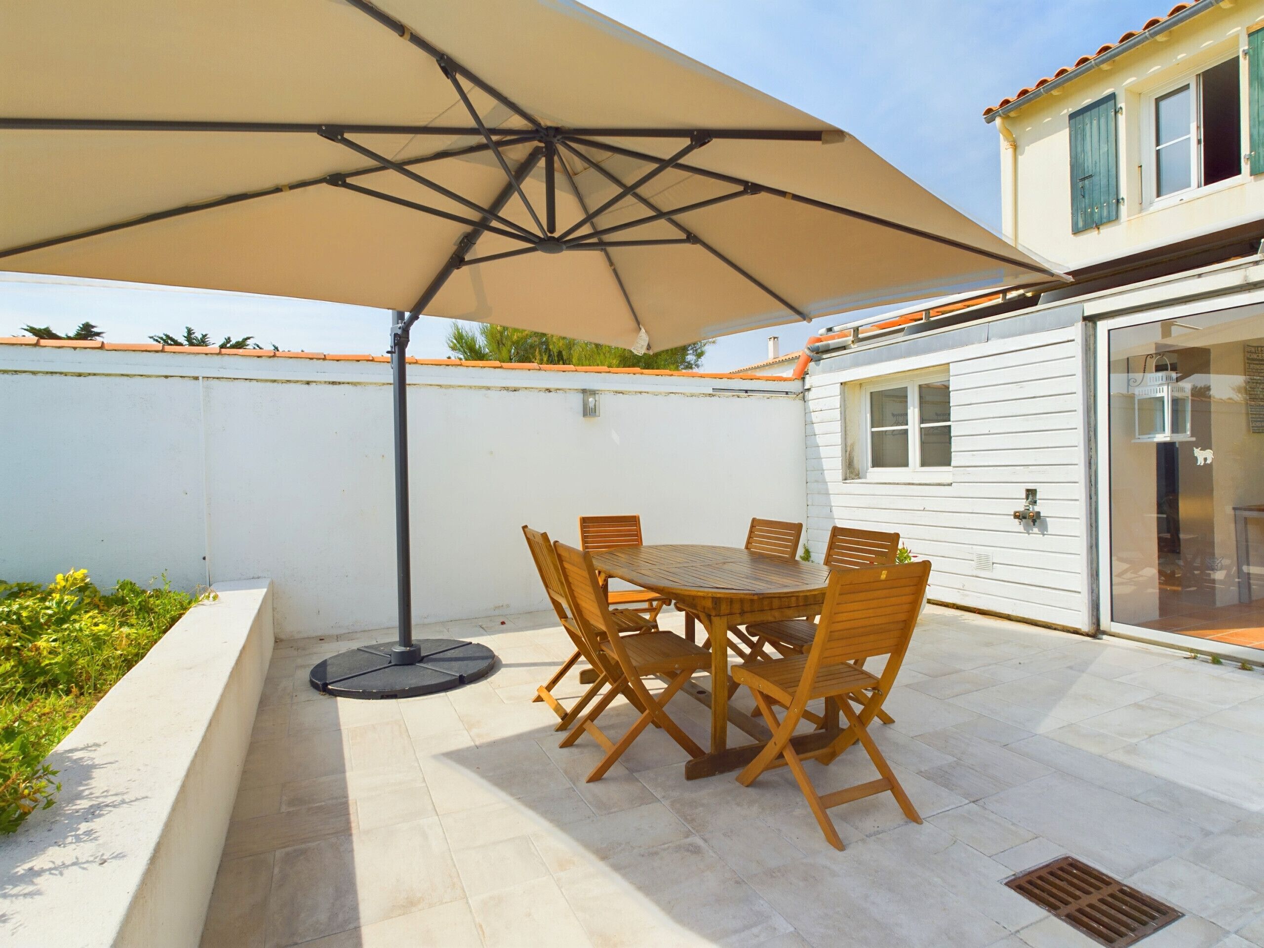 An outdoor patio with a table and chairs under a large umbrella, a white wall, and a building with windows.