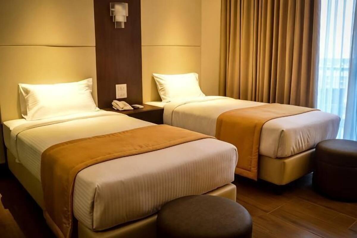 Ein Hotelzimmer mit zwei Betten, einem Nachttisch mit Telefon, einem braunen Hocker und Blick auf die Stadt durch das Fenster.