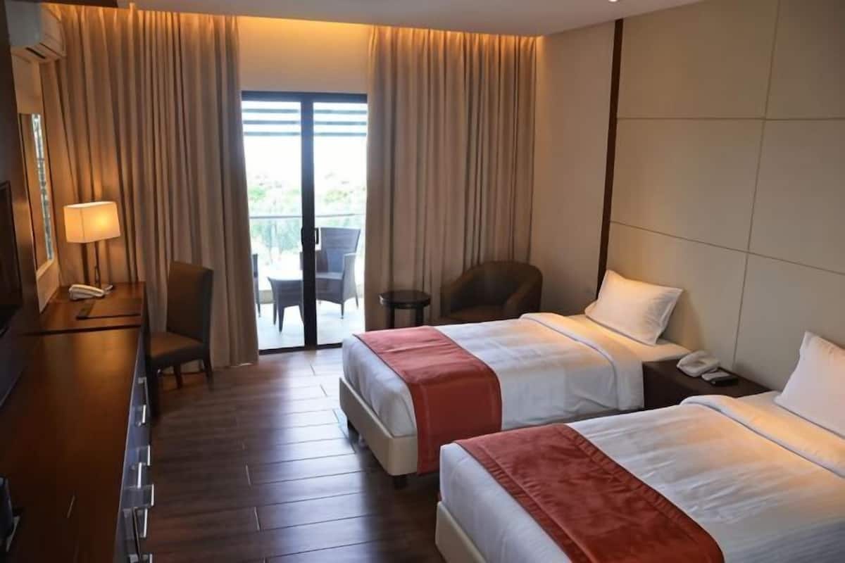 Ein Hotelzimmer mit zwei Betten, einem Schreibtisch mit Lampe, einem Stuhl und Blick ins Freie durch eine Schiebetür.