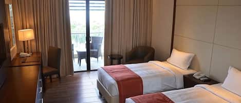 A hotel room with two beds, a desk with a lamp, a chair, and a view of the outdoors through a sliding door.