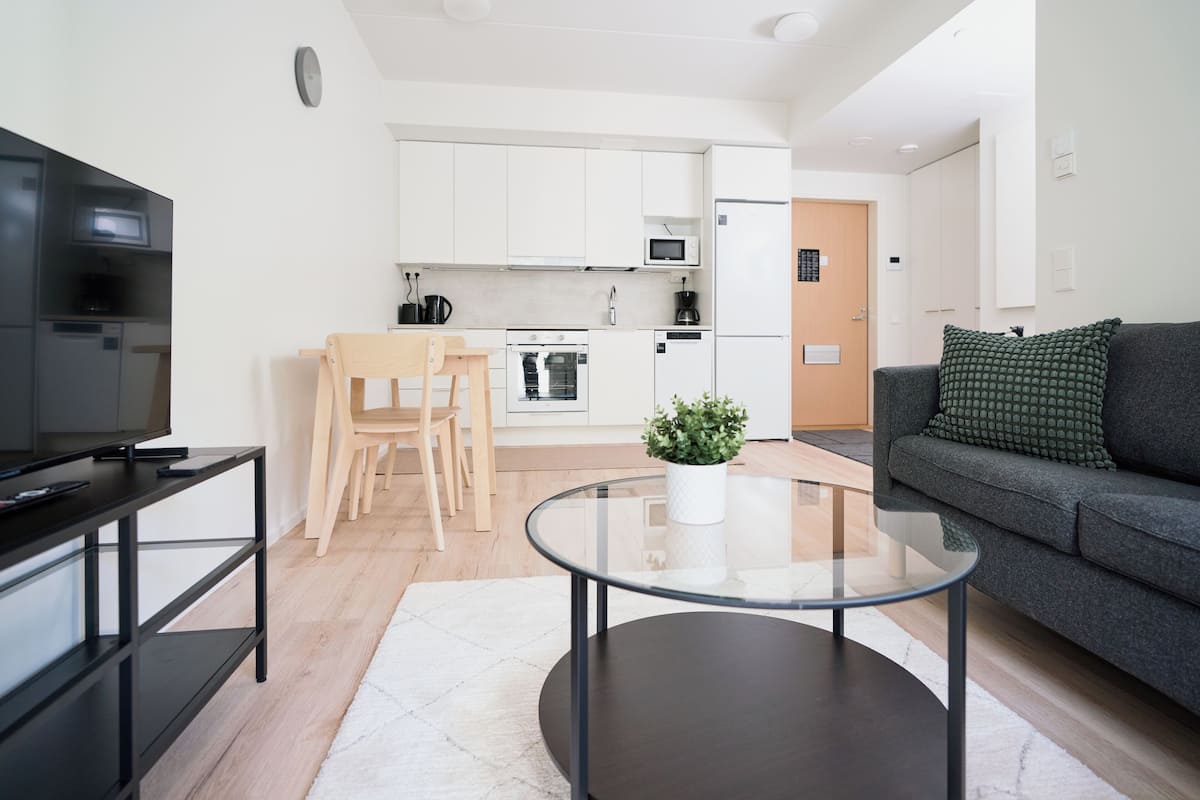 Una sala de estar moderna con una mesa de centro de vidrio, un sofá gris, un televisor sobre un soporte negro y una cocina al fondo.