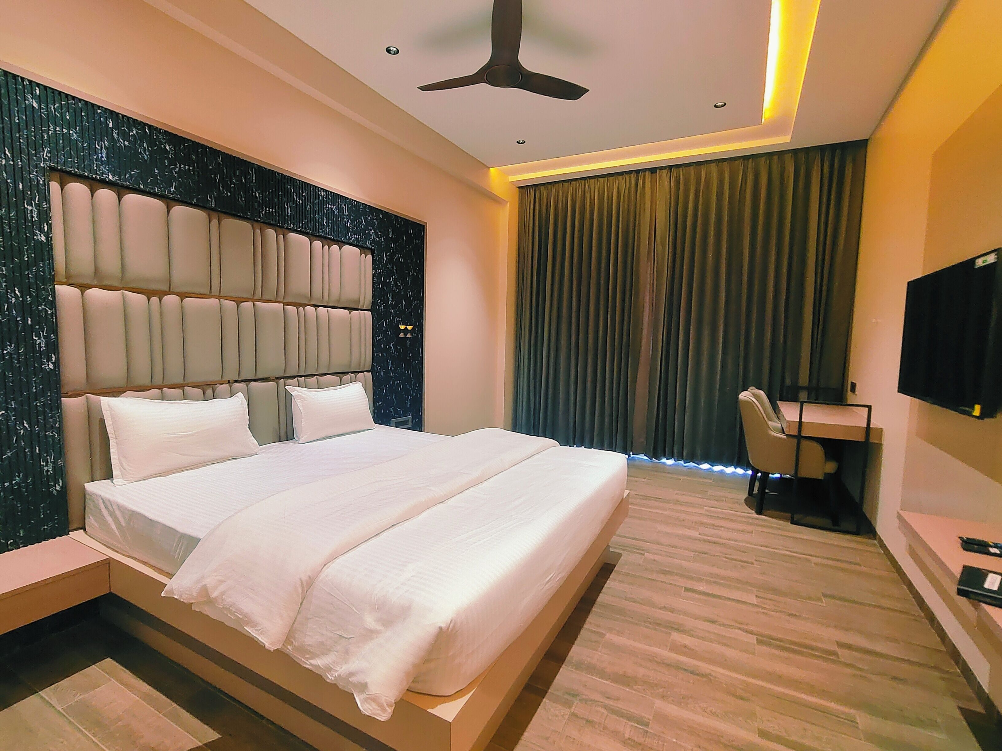 A modern hotel room with a large bed, a desk, a chair, a ceiling fan, and a TV mounted on the wall.
