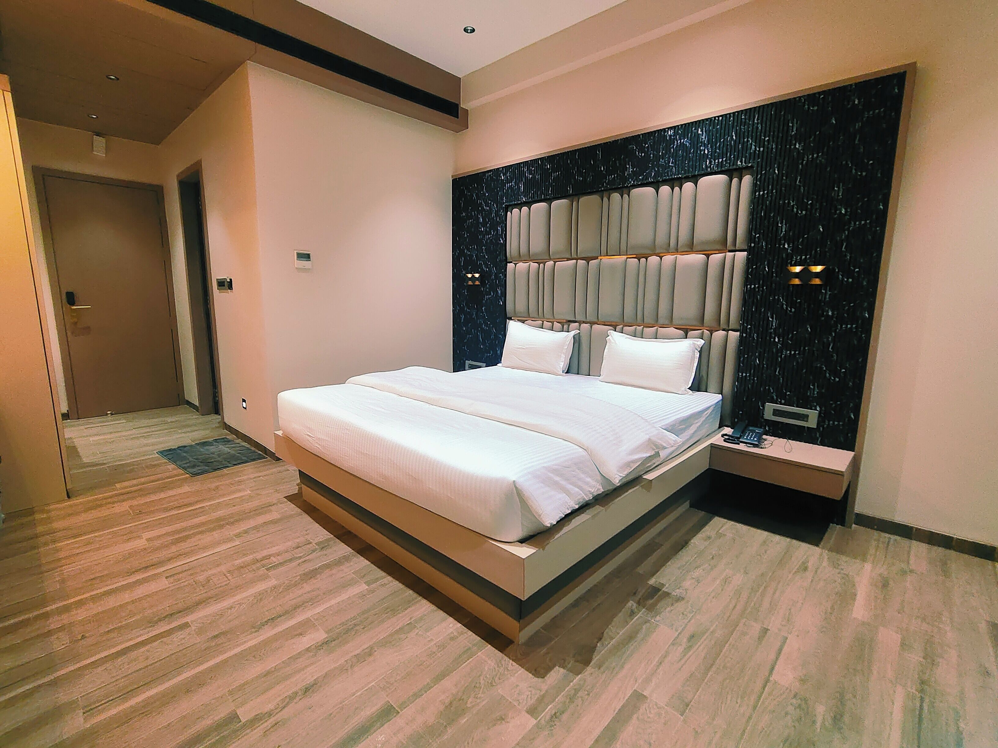 Een moderne hotelkamer met een groot bed, een hoofdbord met verticale panelen, een nachtkastje met een lamp en een vloerbedekking.