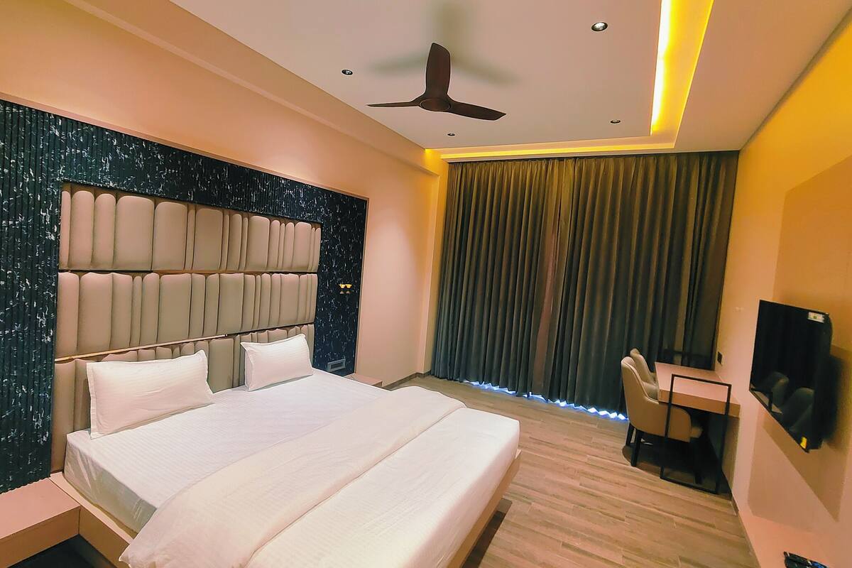 A modern hotel room with a large bed, a desk, a ceiling fan, and a view of a cityscape through the window.