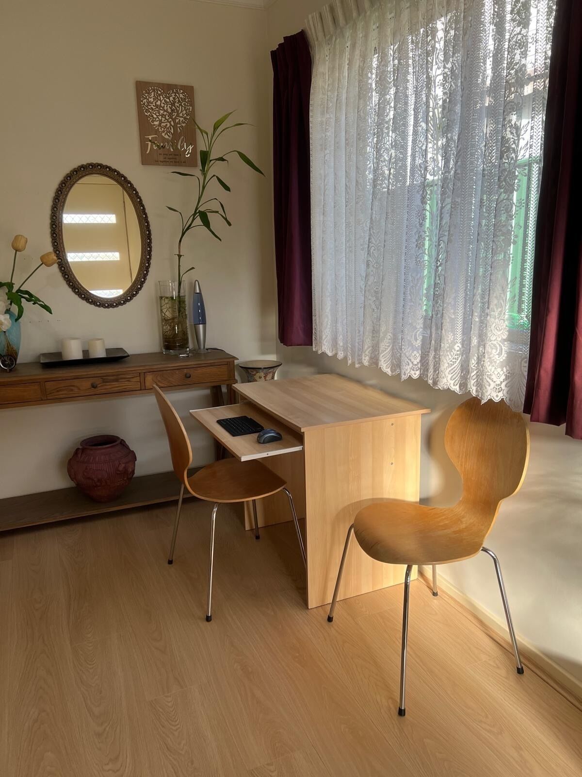 A modern bedroom with a wooden desk, two chairs, a large mirror, a potted plant, and a window with sheer curtains.