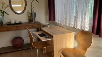 A modern bedroom with a wooden desk, two chairs, a large mirror, a potted plant, and a window with sheer curtains.