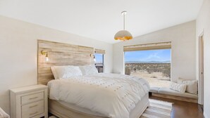 A bedroom with a large bed, bedside tables, a bench with cushions, a view of the desert, and a hanging light fixture. - Desert Moon - Serene Desert Views & Design 3 Bedroom Home by RedAwning (Joshua Tree)