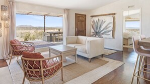 A spacious living room with a large window overlooking a desert landscape, a wooden door, and a large abstract painting on the wall. - Desert Moon - Serene Desert Views & Design 3 Bedroom Home by RedAwning (Joshua Tree)
