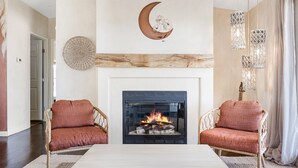 A living room with a fireplace, two wicker chairs with cushions, a coffee table, and a wall-mounted crescent moon decoration. - Desert Moon - Serene Desert Views & Design 3 Bedroom Home by RedAwning (Joshua Tree)