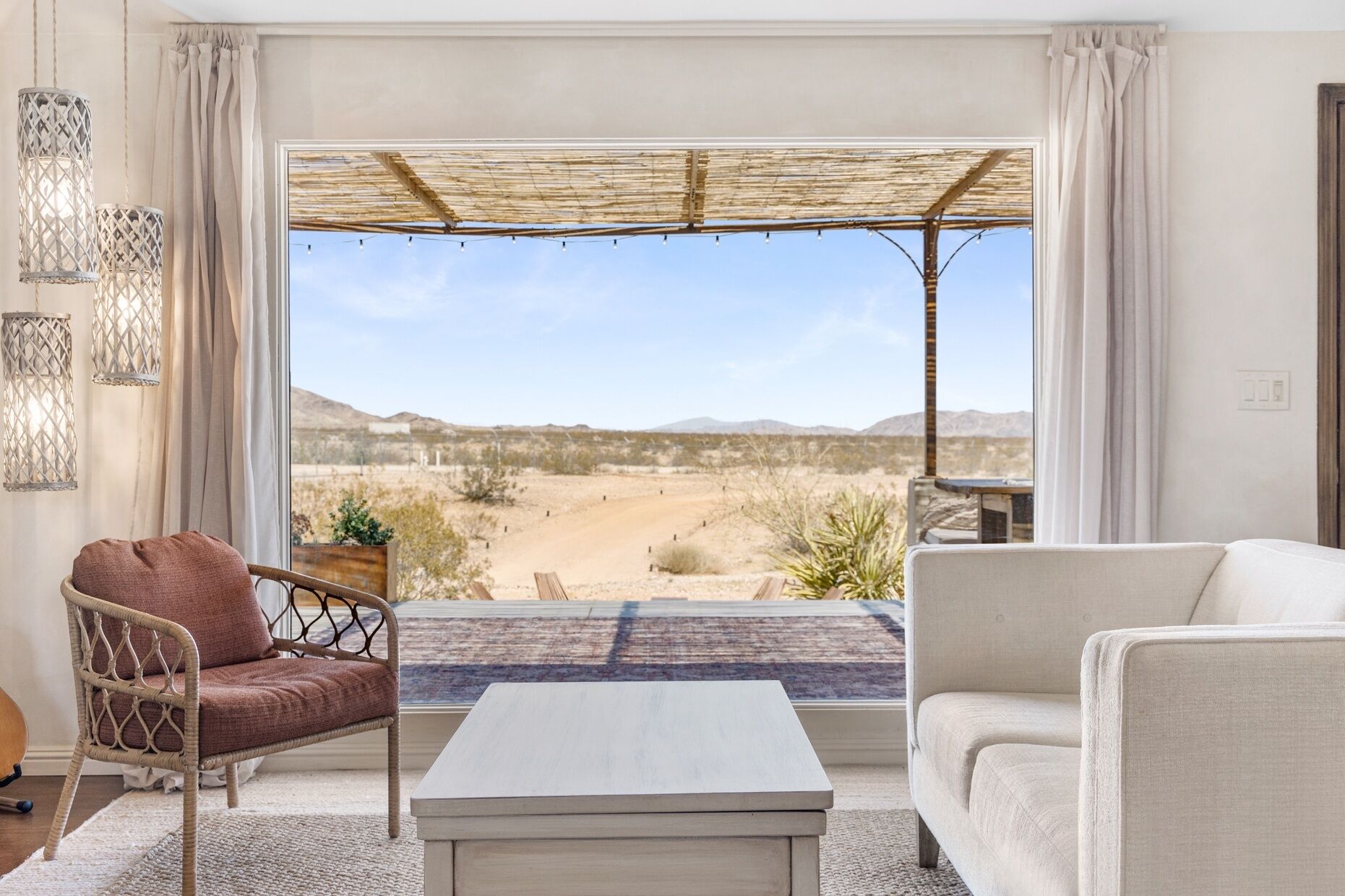 Habitación con vista a un paisaje desértico, que incluye una silla de mimbre, un sofá blanco y una mesa de centro de madera.