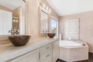 A bathroom with a large mirror, two sinks with vessel basins, a bathtub, and a macramé wall hanging.