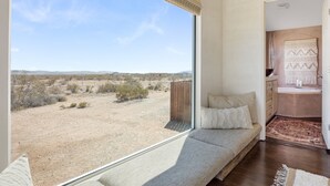 A room with a large window offering a view of a desert landscape. There is a sofa with cushions and a small rug on the floor. - Desert Moon - Serene Desert Views & Design 3 Bedroom Home by RedAwning (Joshua Tree)