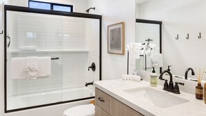 A modern bathroom with a glass shower enclosure, a white toilet, a sink with a marble countertop, and a large mirror. - Yucca Mesa House - Modern Desert Chic 3 Bedroom Home by RedAwning (Yucca Valley)