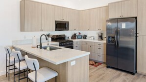 A modern kitchen with light wood cabinets, a stainless steel refrigerator, a built-in oven, and a microwave. There are three bar stools and a sink with a faucet. - Yucca Mesa House - Modern Desert Chic 3 Bedroom Home by RedAwning (Yucca Valley)