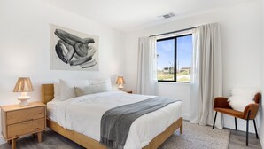 A bedroom with a large bed, two bedside tables with lamps, a chair, a window with curtains, and a painting on the wall. - Yucca Mesa House - Modern Desert Chic 3 Bedroom Home by RedAwning (Yucca Valley)