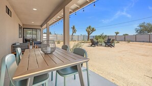 An outdoor patio with a wooden table and chairs, a hanging light fixture, and a view of a desert landscape with cacti and a fence. - Yucca Mesa House - Modern Desert Chic 3 Bedroom Home by RedAwning (Yucca Valley)