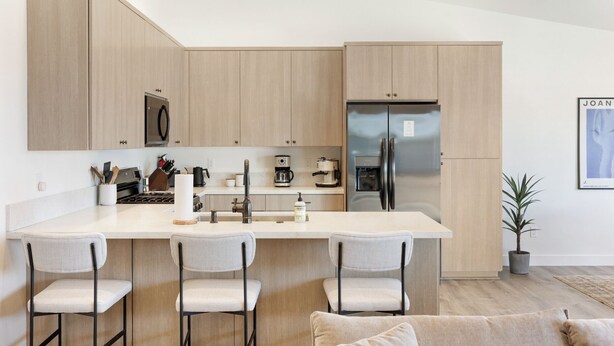 A modern kitchen with wooden cabinets, a stainless steel refrigerator, a coffee machine, and a bar stool area. - Yucca Mesa House - Modern Desert Chic 3 Bedroom Home by RedAwning (Yucca Valley)