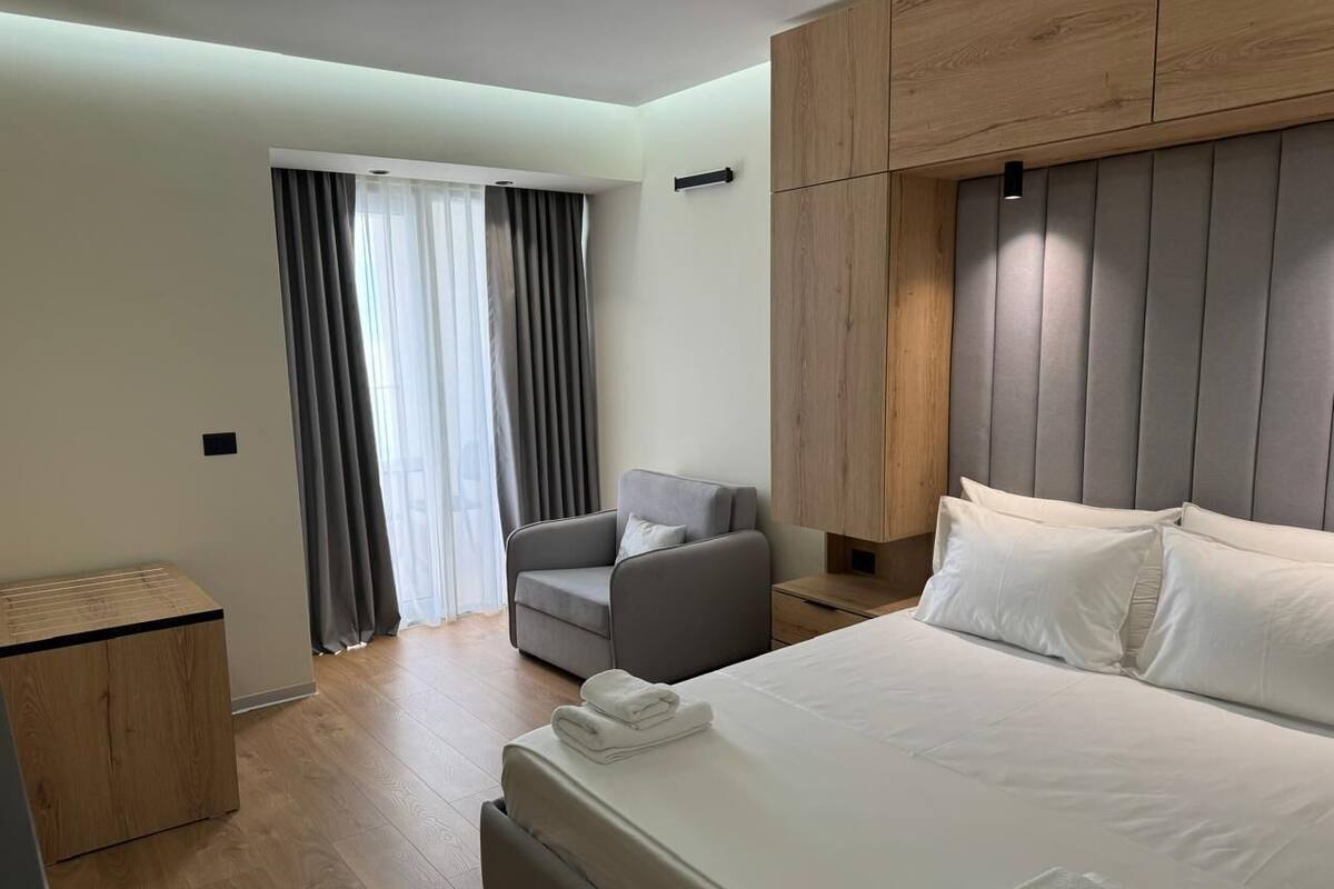 Ein modernes Hotelzimmer mit einem großen Bett, einem grauen Sessel, einem hölzernen Nachttisch und einem Holzschrank.