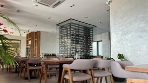 A modern dining area with wooden tables and chairs, a glass display of wine bottles, and a large plant in the foreground. - Sea Side (Durrës)
