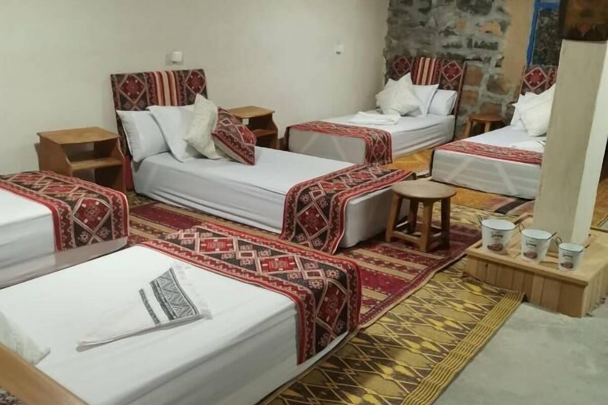 A hotel room with three beds, each with patterned bedding and headboards. There are wooden nightstands and a small table with a chair. The room has a stone wall and a mirror.