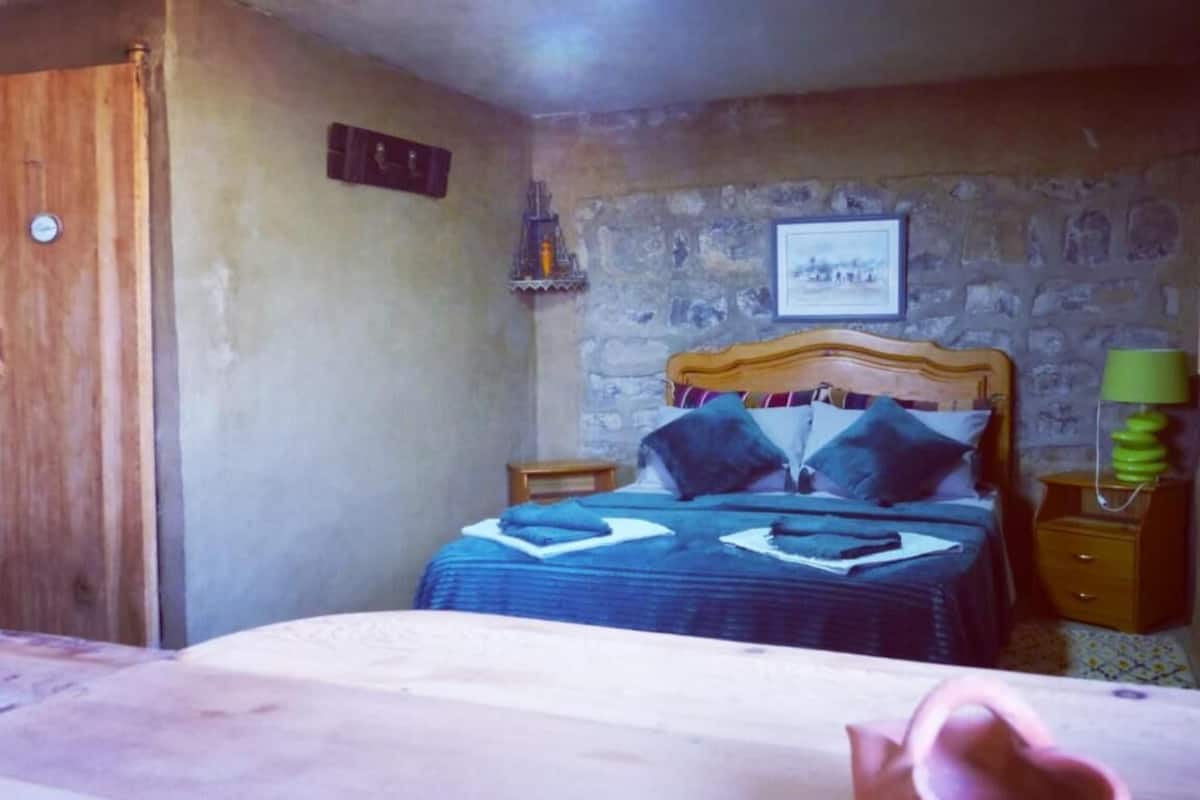 A bedroom with a wooden bed, a stone wall, a small bedside table, and a nightstand with a lamp.