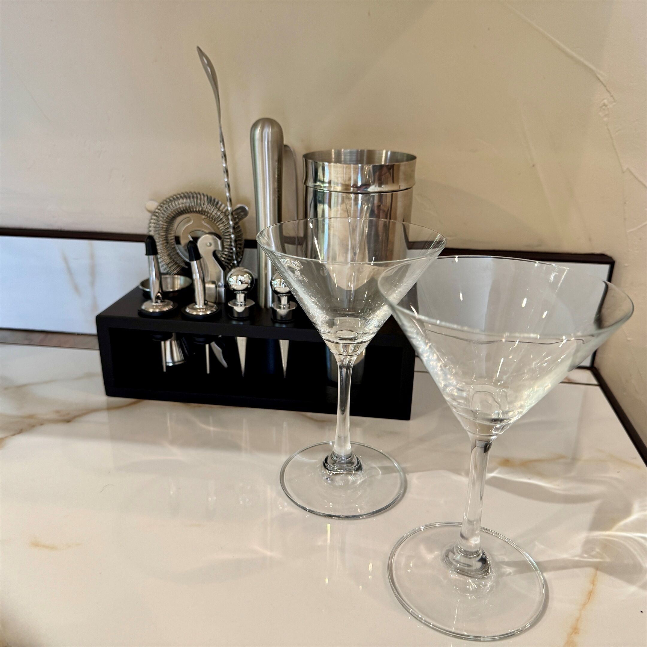 Two martini glasses on a marble countertop with a black tray holding various kitchen utensils.
