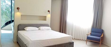 Quarto de hotel moderno com uma cama grande, uma cadeira, um ventilador de teto e vista para o verde lá fora.