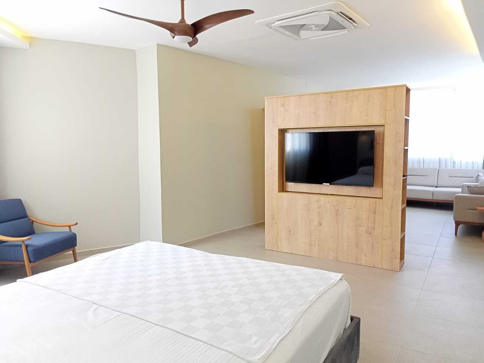 Une chambre d’hôtel moderne équipée d’un lit, d’un téléviseur fixé sur un panneau en bois, d’un ventilateur de plafond et d’un coin salon avec un canapé et des fauteuils.