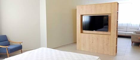 A modern hotel room with a bed, a TV mounted on a wooden panel, a ceiling fan, and a seating area with a sofa and chairs.