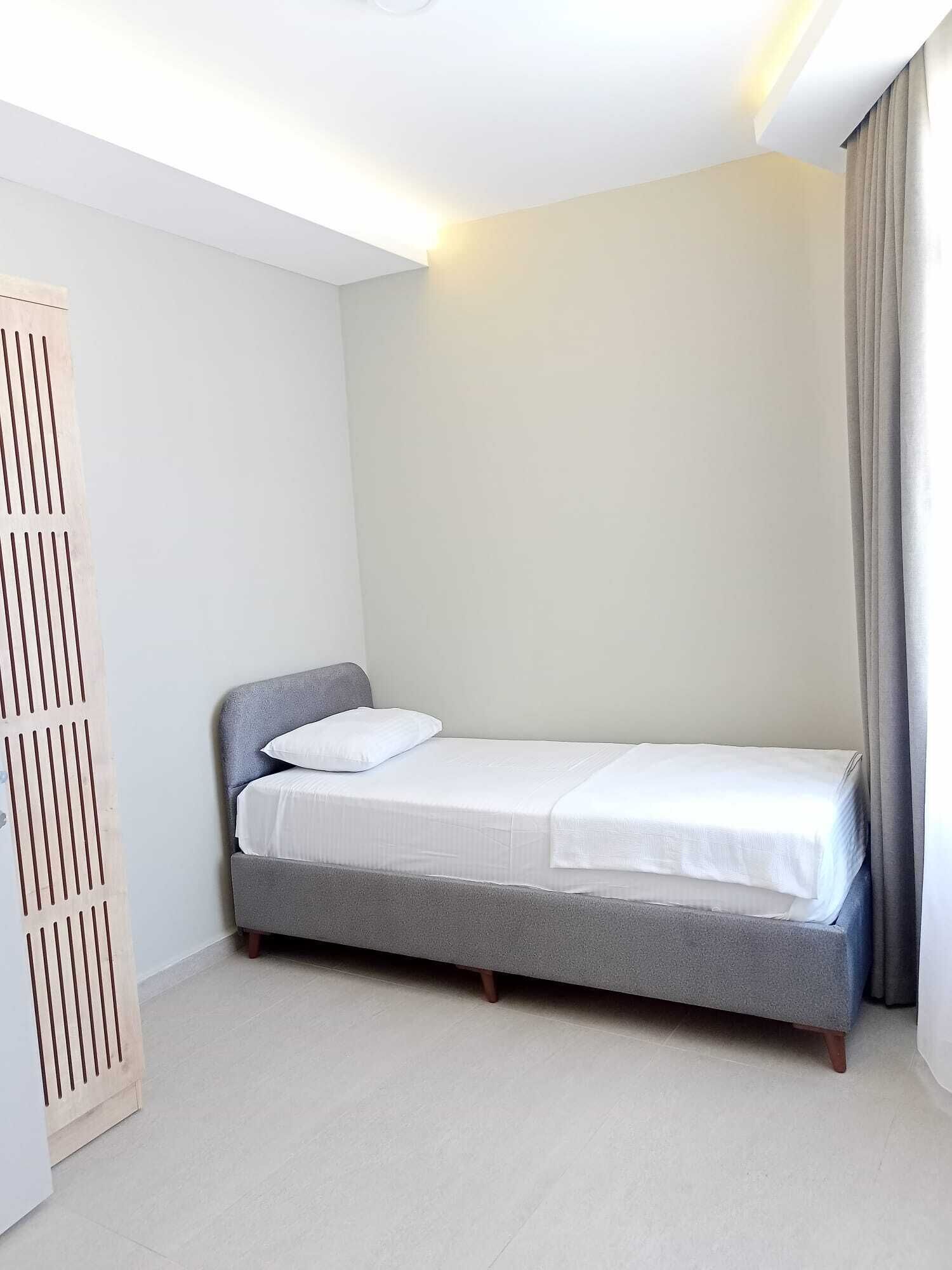 A single bed with a grey headboard and white bedding in a room with white walls and a light-colored floor.