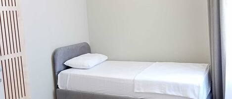 A single bed with a grey headboard and white bedding in a room with white walls and a light-colored floor.
