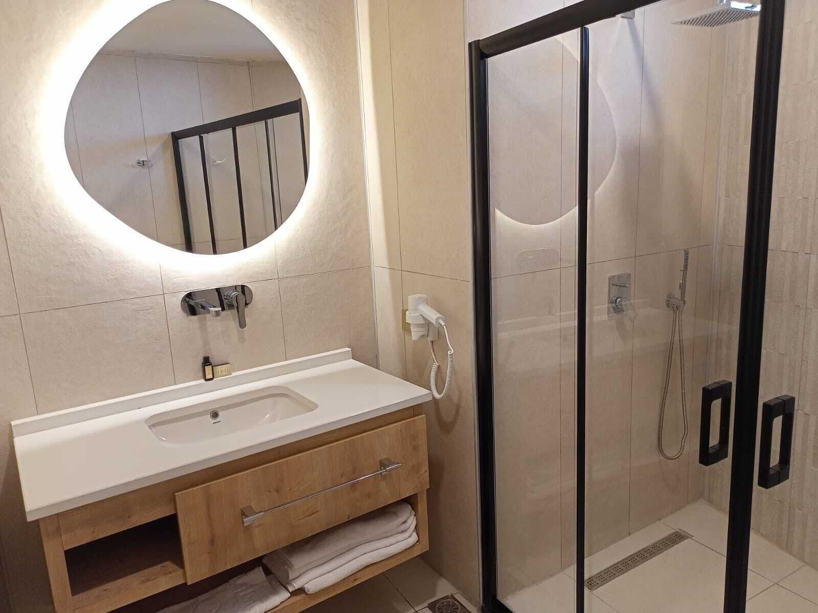 Un baño moderno con lavamanos blanco, tocador de madera, cabina de ducha de vidrio y espejo redondo con luz.